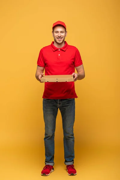 Full length view of happy delivery man in red uniform holding pizza box on yellow — Stock Photo