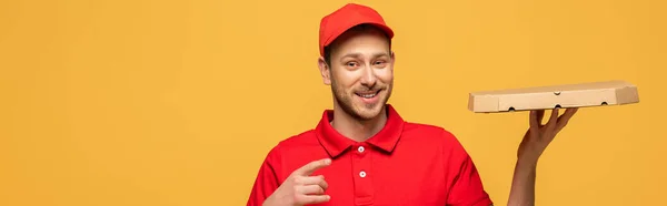 Feliz repartidor en uniforme rojo apuntando con el dedo a la caja de pizza aislado en amarillo, tiro panorámico — Stock Photo