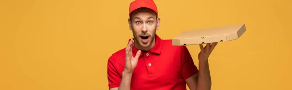 Felice fattorino in uniforme rossa che tiene la scatola della pizza a bocca aperta isolata sul giallo, colpo panoramico — Foto stock