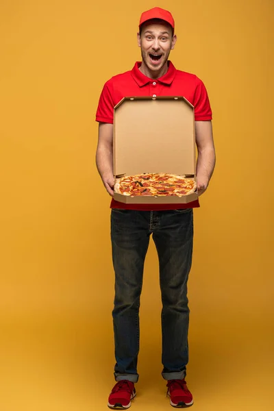 Happy delivery man with open mouth in red uniform showing box with delicious pizza on yellow — Stock Photo