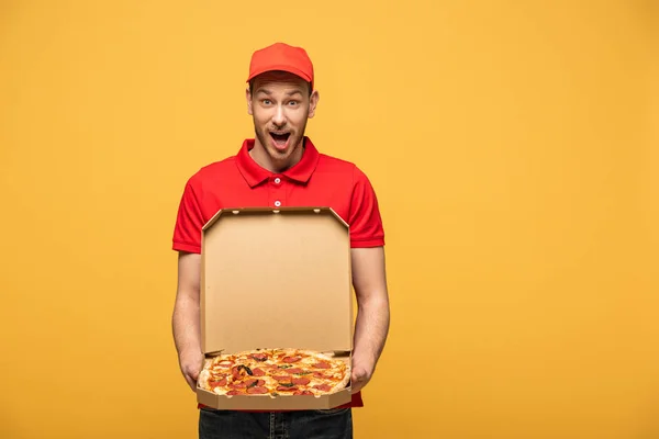 Sorprendido repartidor hombre en uniforme rojo mostrando caja con deliciosa pizza aislada en amarillo - foto de stock