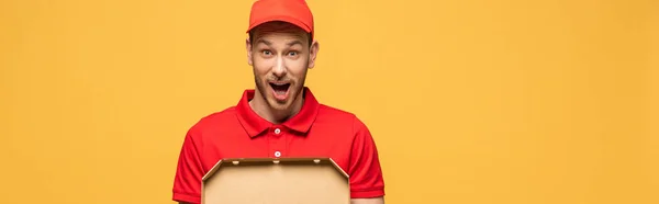 Chocado repartidor en uniforme rojo sosteniendo caja de pizza aislado en amarillo, tiro panorámico - foto de stock