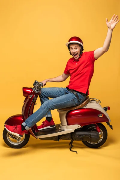 Vista lateral del repartidor en scooter agitando la mano sobre fondo amarillo - foto de stock