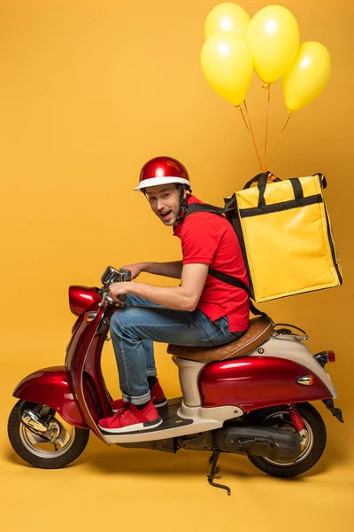 Вид збоку щасливого чоловіка доставки з повітряними кулями на рюкзаку на скутері на жовтому фоні — стокове фото