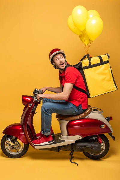 Side view of delivery man looking at balloons on backpack on scooter on yellow background — Stock Photo