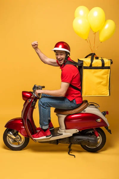 Side view of happy delivery man with balloons on backpack on scooter showing fist on yellow background — Stock Photo