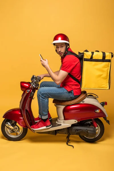 Вид сбоку доставщика с рюкзаком на скутере со смартфоном на желтом фоне — стоковое фото