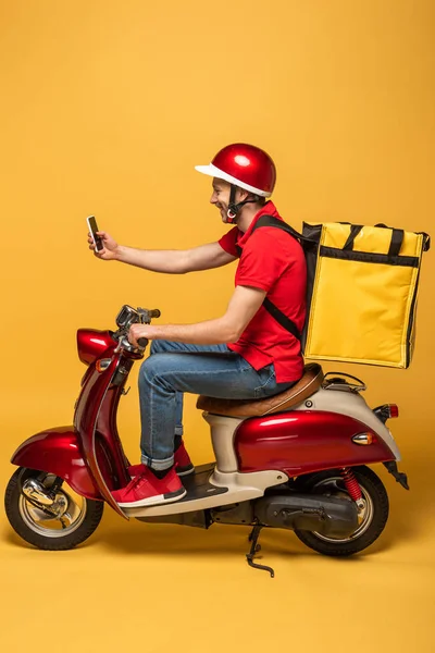 Vista lateral do homem de entrega com mochila em scooter tomando selfie no smartphone no fundo amarelo — Fotografia de Stock