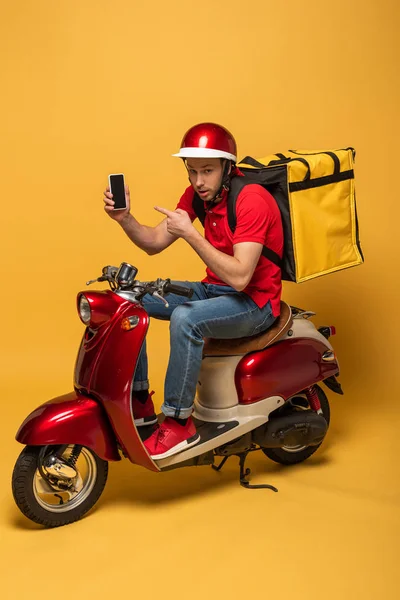 Repartidor con mochila en scooter apuntando al teléfono inteligente sobre fondo amarillo - foto de stock