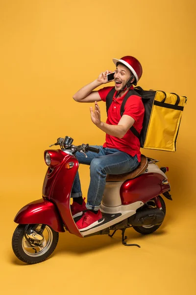Happy delivery man with backpack on scooter talking on smartphone on yellow background — Stock Photo