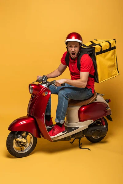 Delivery man with backpack on scooter screaming on yellow background — Stock Photo