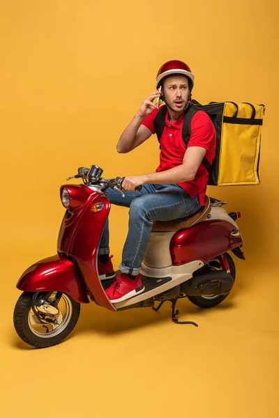 Homem de entrega preocupado com mochila em scooter falando no smartphone no fundo amarelo — Fotografia de Stock