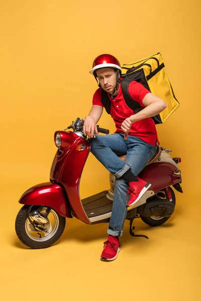 Sad delivery man with backpack on scooter showing thumb down on yellow background — Stock Photo