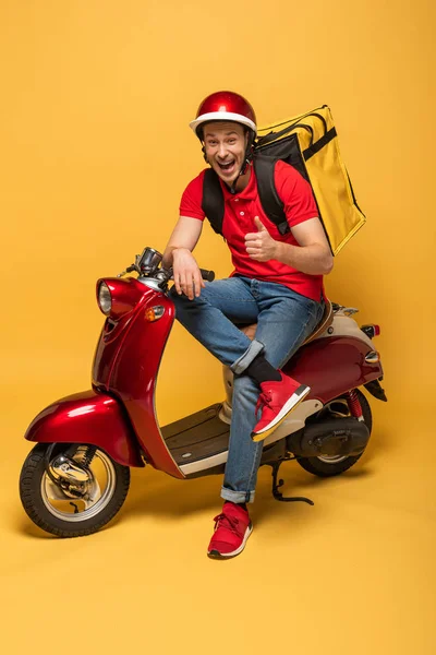 Happy delivery man with backpack on scooter showing thumb up on yellow background — Stock Photo