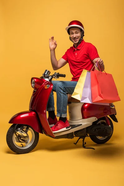 Livreur souriant en uniforme rouge avec des sacs à provisions agitant la main sur scooter sur fond jaune — Photo de stock