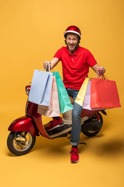 Repartidor sonriente en uniforme rojo con bolsas de compras en scooter sobre fondo amarillo - foto de stock