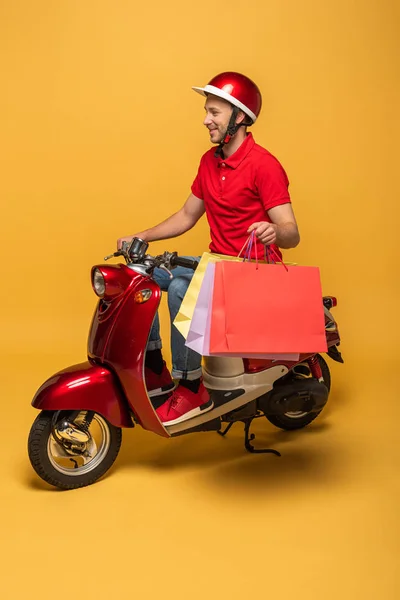 Lächelnder Zusteller in roter Uniform mit Einkaufstaschen auf Roller auf gelbem Hintergrund — Stockfoto