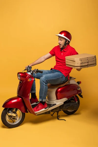 Homem de entrega chocado em uniforme vermelho segurando caixas de pizza em scooter no fundo amarelo — Fotografia de Stock