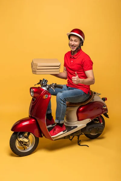 Homem de entrega sorrindo em vermelho uniforme segurando caixas de pizza e mostrando polegar para cima em scooter no fundo amarelo — Fotografia de Stock