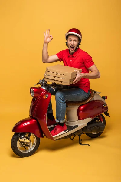 Excited delivery man in red uniform holding pizza boxes and showing ok sign on scooter on yellow background — Stock Photo