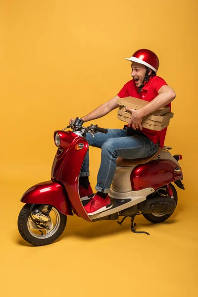 Repartidor excitado en uniforme rojo sosteniendo cajas de pizza en scooter sobre fondo amarillo - foto de stock