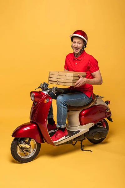 Hombre de entrega feliz en uniforme rojo sosteniendo cajas de pizza en scooter sobre fondo amarillo - foto de stock