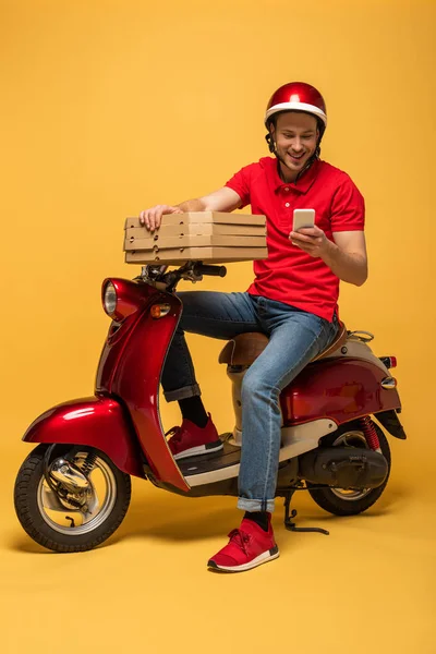 Felice uomo di consegna in uniforme rossa in possesso di scatole di pizza e utilizzando smartphone su scooter su sfondo giallo — Foto stock