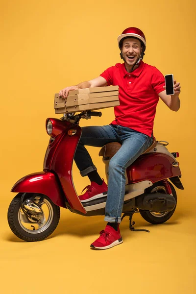 Heureux livreur en uniforme rouge tenant boîtes à pizza et smartphone avec écran blanc sur scooter sur fond jaune — Photo de stock