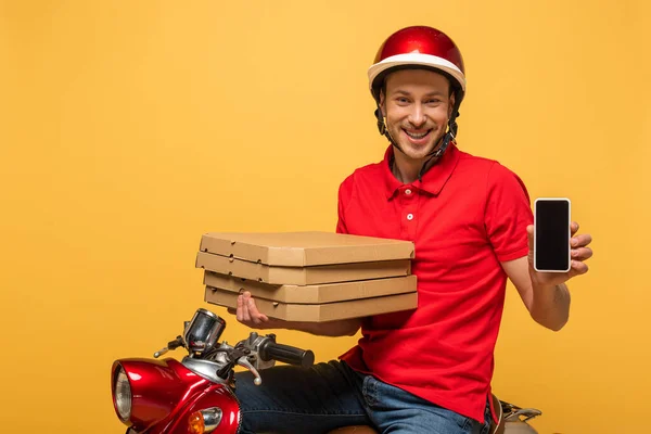 Hombre de entrega feliz en uniforme rojo sosteniendo cajas de pizza y teléfono inteligente con pantalla en blanco en scooter aislado en amarillo - foto de stock