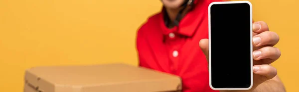 Vista recortada del hombre de entrega feliz en uniforme rojo sosteniendo cajas de pizza y teléfono inteligente con pantalla en blanco aislado en amarillo, tiro panorámico - foto de stock