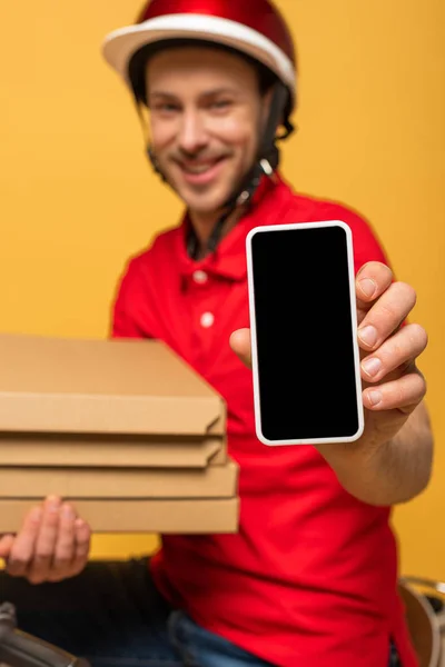 Messa a fuoco selettiva di felice consegna uomo in uniforme rossa contenente scatole di pizza e smartphone con schermo bianco isolato su giallo — Foto stock