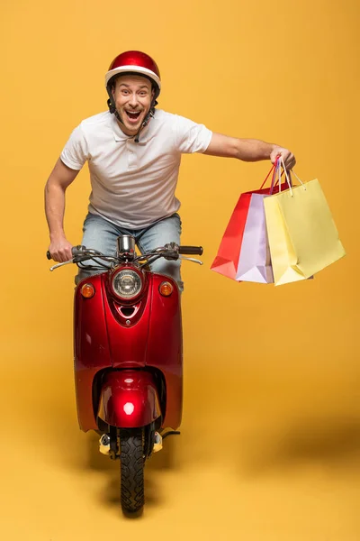 Happy delivery man in helmet riding scooter with shopping bags on yellow background — Stock Photo