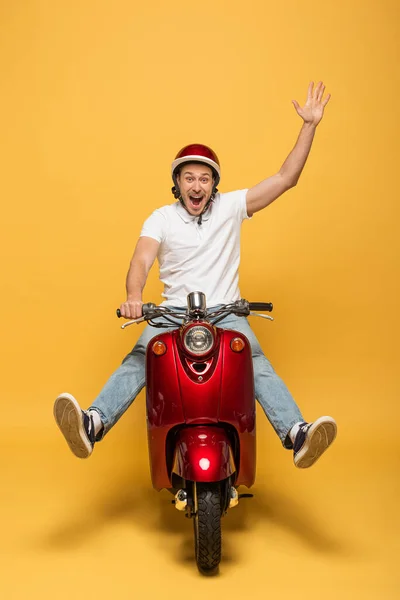 Happy delivery man in helmet riding scooter with raised hand on yellow background — Stock Photo
