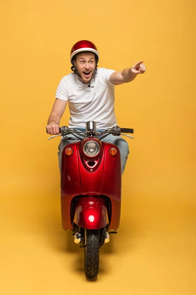 Щасливий чоловік доставки в шолом їде скутер і вказує пальцем на жовтий фон — стокове фото