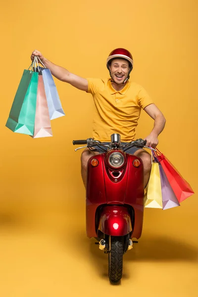 Heureux livreur en uniforme jaune scooter équitation avec des sacs à provisions sur fond jaune — Photo de stock