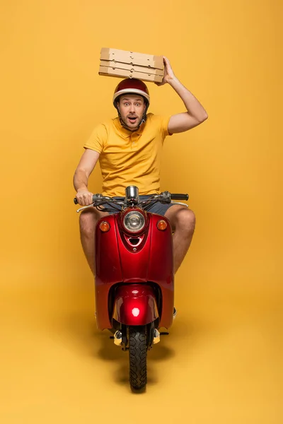 Schockierter Zusteller in gelber Uniform fährt Roller mit Pizzakartons auf gelbem Hintergrund — Stockfoto