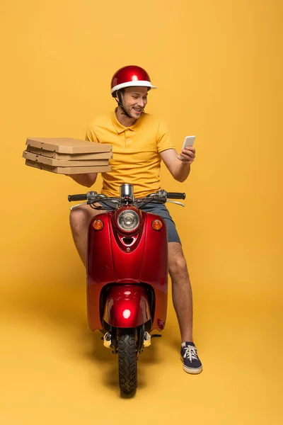 Repartidor sonriente en scooter uniforme amarillo con cajas de pizza y uso de teléfono inteligente sobre fondo amarillo - foto de stock