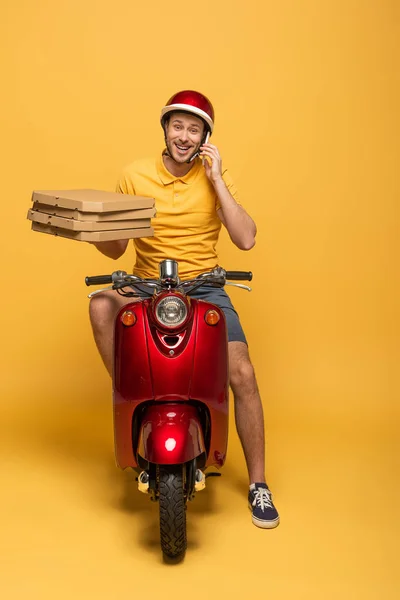 Repartidor sonriente en scooter uniforme amarillo con cajas de pizza y hablando en smartphone sobre fondo amarillo - foto de stock