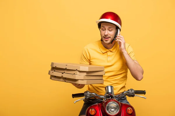 Homem de entrega em scooter de equitação uniforme amarelo com caixas de pizza e falando no smartphone isolado no amarelo — Fotografia de Stock