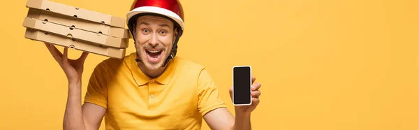 Repartidor excitado en uniforme amarillo con cajas de pizza mostrando teléfono inteligente con pantalla en blanco aislado en amarillo, tiro panorámico - foto de stock