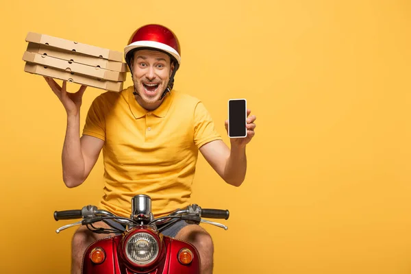 Repartidor excitado en scooter uniforme amarillo con cajas de pizza y mostrando teléfono inteligente con pantalla en blanco aislado en amarillo - foto de stock