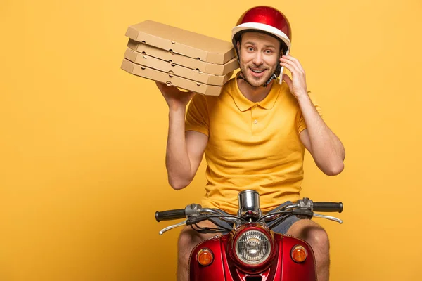 Smiling delivery man in yellow uniform riding scooter with pizza boxes and talking on smartphone isolated on yellow — Stock Photo