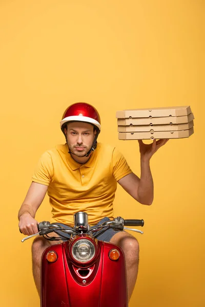 Repartidor concentrado en scooter uniforme amarillo con cajas de pizza aisladas en amarillo - foto de stock