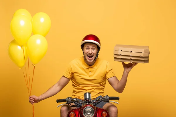 Happy delivery man in yellow uniform riding scooter with pizza boxes and balloons isolated on yellow — Stock Photo