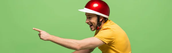Vista lateral del hombre feliz entrega en uniforme amarillo y casco apuntando con el dedo aislado en verde, tiro panorámico — Stock Photo