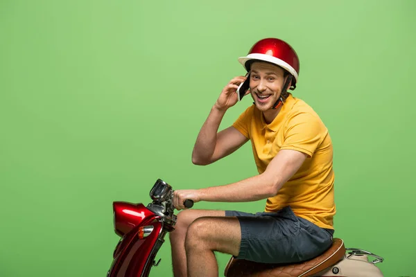 Seitenansicht eines glücklichen Zustellers in gelber Uniform, der auf dem Smartphone auf einem Roller auf Grün redet — Stockfoto