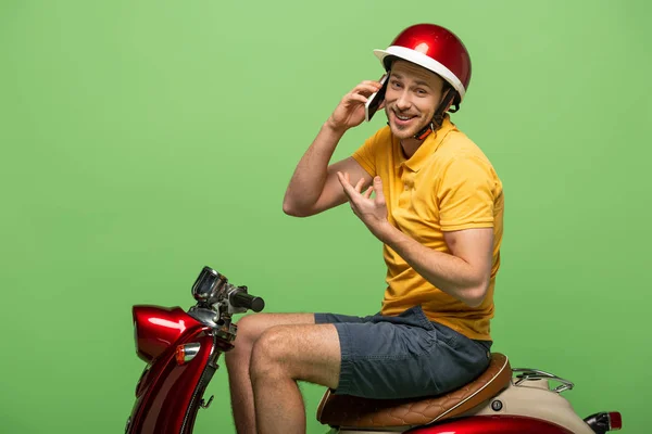 Visão lateral do homem de entrega feliz em uniforme amarelo falando no smartphone em scooter isolado no verde — Fotografia de Stock