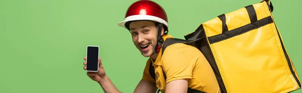 Side view of happy delivery man in yellow uniform with backpack showing smartphone with blank screen isolated on green, panoramic shot — Stock Photo