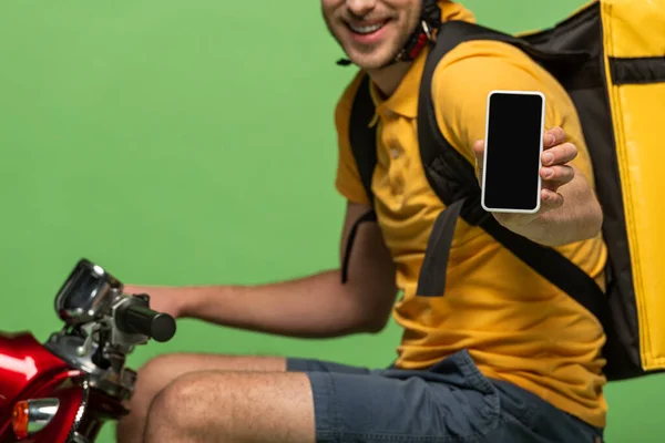 Cropped view of happy delivery man in yellow uniform with backpack showing smartphone with blank screen on scooter isolated on green — Stock Photo