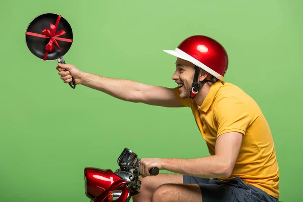 Side view of happy delivery man in yellow uniform holding frying pan with bow on scooter isolated on green — Stock Photo